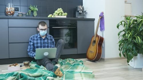 Man in medical face mask working on laptop at remote workplace at home kitchen