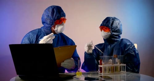 Dos personas con ropa protectora, respiradores, gafas y guantes están sentadas en una mesa.
Ellos