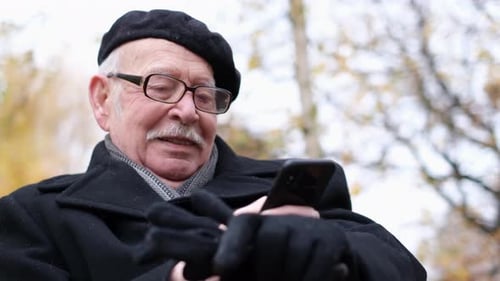 The Pensioner Uses a Smartphone He is Sitting on a Park Bench