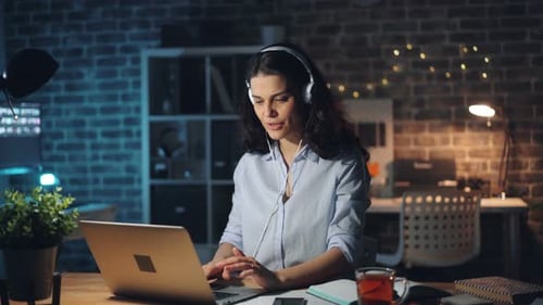 Cute Girl Working with Laptop and Enjoying Music in Headphone in Office at Night