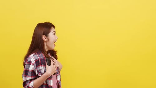 Asian female using phone with positive expression, smiles broadly over yellow background