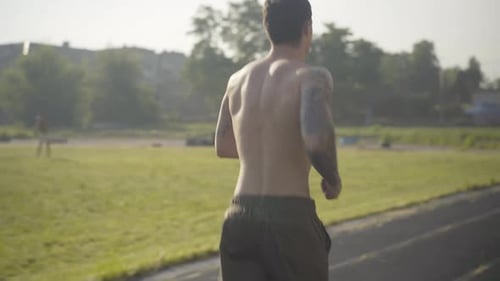 Back View of Sportive Caucasian Man Jogging on Stadium in Summer Morning. Camera Follows Young