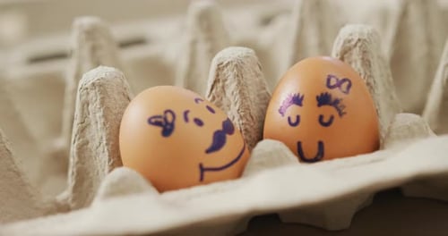 Video of close up of two brown eggs with drawn faces in egg carton background