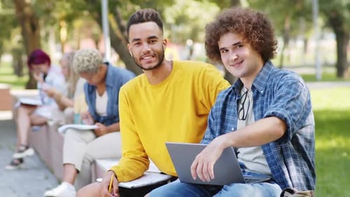 Portrait of Two Cheerful Male Students Sitting with Laptop in Park