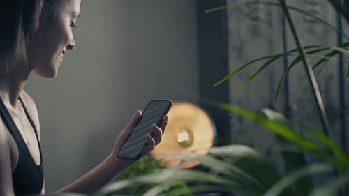 Side View of Young Fitness and Brunette Woman Smiles Looking at the Mobile During a Workout Rest in
