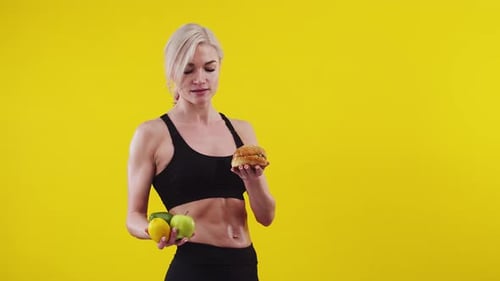 Mujer atlética sostiene en las manos y elige una fruta o hamburguesa en el estudio un fondo naranja