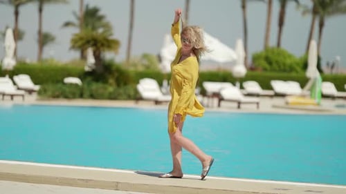 a Curly Blonde in Sunglasses and a Yellow Fluttering Dress Walks Along the Edge of the Pool on a
