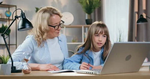 Madre con gafas ayudando a su inteligente hija con los deberes, sentada en el lugar de trabajo