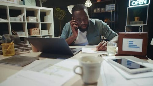 Portrait of Busy Afro Manager Writing Notes on Documents Near Laptop in Office.