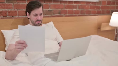 Man Working from Bed with Laptop and Document