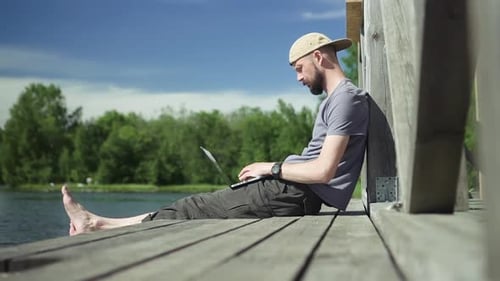 Guy with Laptop Sit on Lake Pier and Types on Computer Spbd