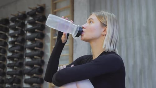 fitness girl drinking from sport bottle during a workout pause at home or gym. Close backlit