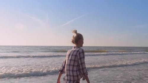 A slender woman walking on the waves of sea water on a sandy beach.
