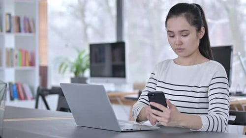 Young Asian Woman with Laptop Using Smartphone in Office
