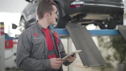 Porträt eines konzentrierten kaukasischen Mannes, der ein Tablet benutzt und in der Werkstatt auf ein Auto zurückblickt