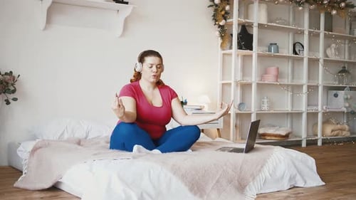 Young Plus Size Woman Watching Online Yoga on Laptop Sitting on Bed and Practicing Meditation in