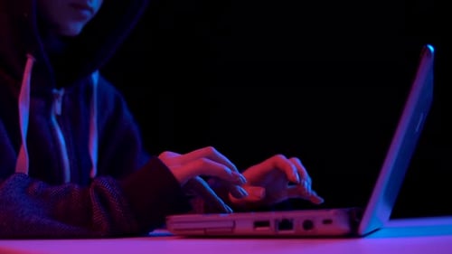 Young Woman in a Hood with a Laptop. Hacker Makes a Hack Through a Laptop. Blue and Red Light Falls