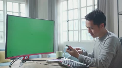 Asian Man With Green Screen Desktop Computer Using Mobile Phone While Working At Home