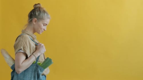 Retrato de mujer joven caminando con bolsa de compras reutilizable con comida sobre fondo amarillo