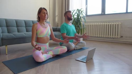 Pareja meditando en casa frente a una computadora portátil con un tutorial de yoga en línea