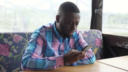 Afro American Man Is Browsing Smartphone Sitting in Cafe Waiting His Order