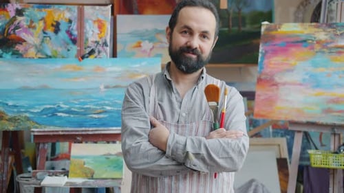 Retrato de um pintor adulto em pé no estúdio de arte segurando pincéis e olhando para a câmera