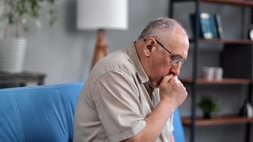 Unwell Gray Haired 70s Elderly Man Coughing with Seasonal Flu Symptoms Sitting on Couch at Home