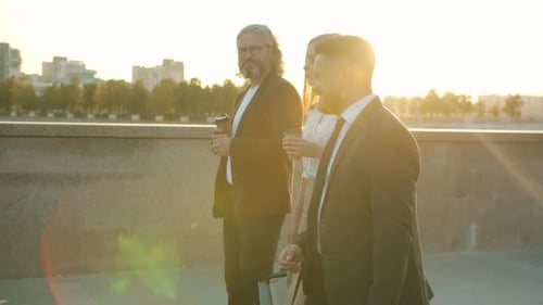 Side View of Cheerful Businesspeople Walking Outdoors in City in Sunny Morning Holding to Go Coffee