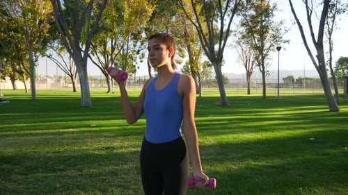 A young woman doing a dumbbell workout in the park performing bicep curls to build arm muscle and st