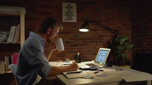 Kaukasischer Mann mit hellem Bart und blauem Hemd trinkt müde von einer weißen Tasse neben dem Computer am Tisch
