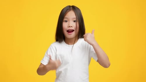Kleines asiatisches Mädchen, das mit dem Daumen nach oben gestikuliert und auf gelbem Hintergrund posiert