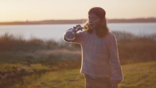 Girl Blowing Bubbles With Wand In Sunlit Field At Sunset