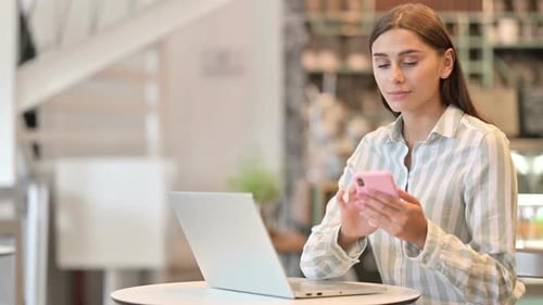 Young Latin Woman Using Smartphone and Laptop in Cafe