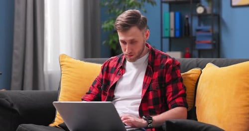 A Handsome College Student Boy is Studying Remotely While Sitting on Couch in Room at