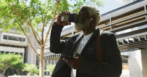 African american senior man drinking coffee and using smartphone in corporate park
