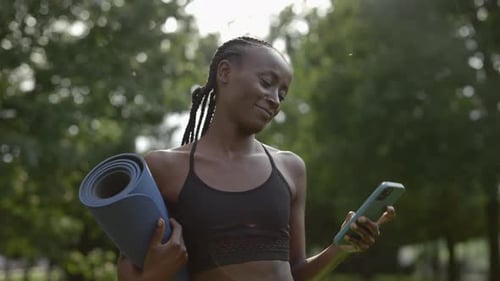 Woman Carrying Yoga Mat and Using Mobile at City Park