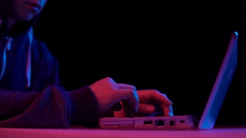 Young Man in a Hood with a Laptop Closeup. Hacker Makes a Hack Through a Laptop. Blue and Red Light