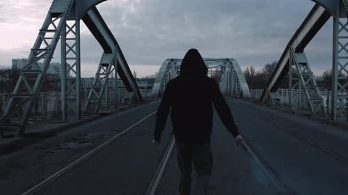 Young Man in Hoodie and Balaclava with Red Burning Signal Flare on the Road Under an Old Steel Frame