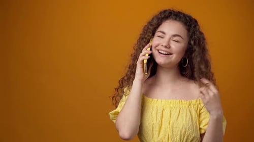 Jeune femme frisée parlant au téléphone et souriant sur fond jaune