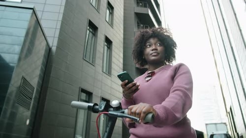 Afro Woman Standing with E-Scooter in City and Using Smartphone