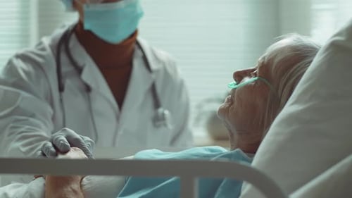 Doctor in Mask Supporting Senior Woman in Hospital Ward