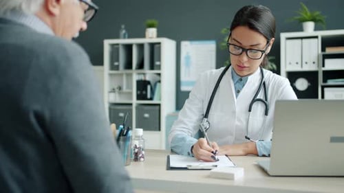 Female Medic Consulting Old Man Patient in Hospital Talking Writing in Chart