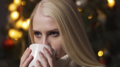 Woman Drinks from Mug by Christmas Tree at Night