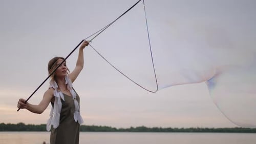 A Young Female Artist Shows a Soap Bubble Show Blowing Up Huge Soap Bubbles on the Shore of a Lake