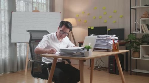 Asian Man Looking At Laptop And Taking Note While Working With Documents At The Office
