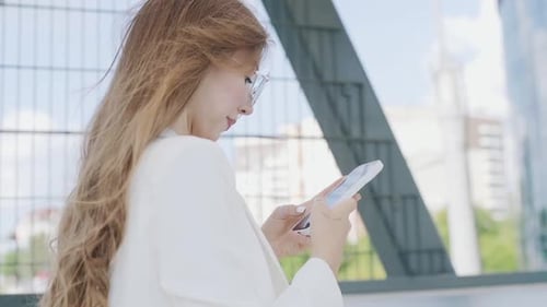 Smiling woman wearing trendy glasses walks down the central city street and uses her phone. Pretty s