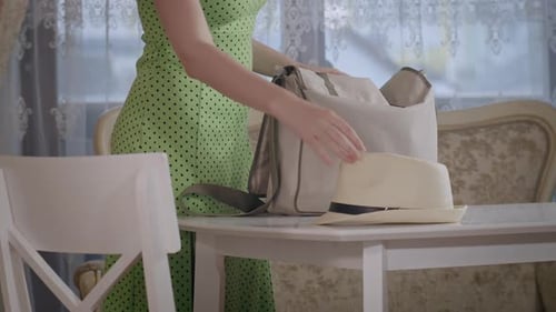 A Girl in a Dress Puts a Bag on the Table and Lays a White Hat Next to It