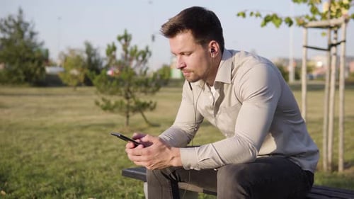 A Young Caucasian Man Listens to Music with Earphones on a Smartphone As He Sits in a Park