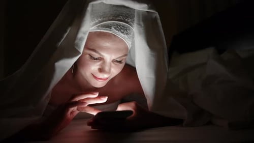 Beautiful Young Woman Using Smart Phone Under Blanket in Bedroom on Bedtime