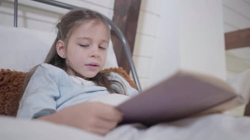 Beautiful Caucasian Girl with Grey Eyes Holding Book and Reading. Portrait of Little Lady Lying in